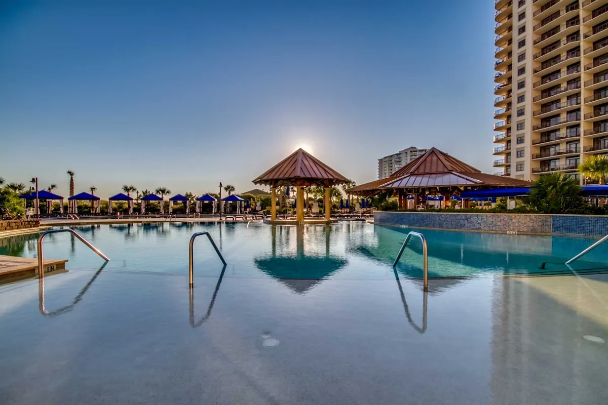 This image features an expansive swimming pool surrounded by thatched-roof cabanas and lounge chairs, all bathed in the warm hues of a sunset. The setting conveys a sense of relaxation and resort-style amenities, perfect for family fun, swimming, and socializing in a picturesque environment.