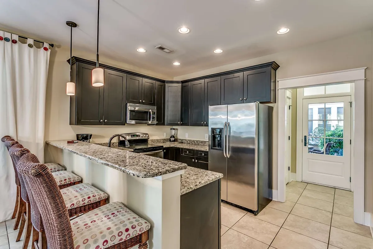 A kitchen scene featuring a granite countertop with bar stools and an array of kitchen appliances. The entrance door is visible, contributing to the sense of openness and easy access between the kitchen and other areas. The mood is welcoming, suggesting socializing and entertaining around this hub of the home.