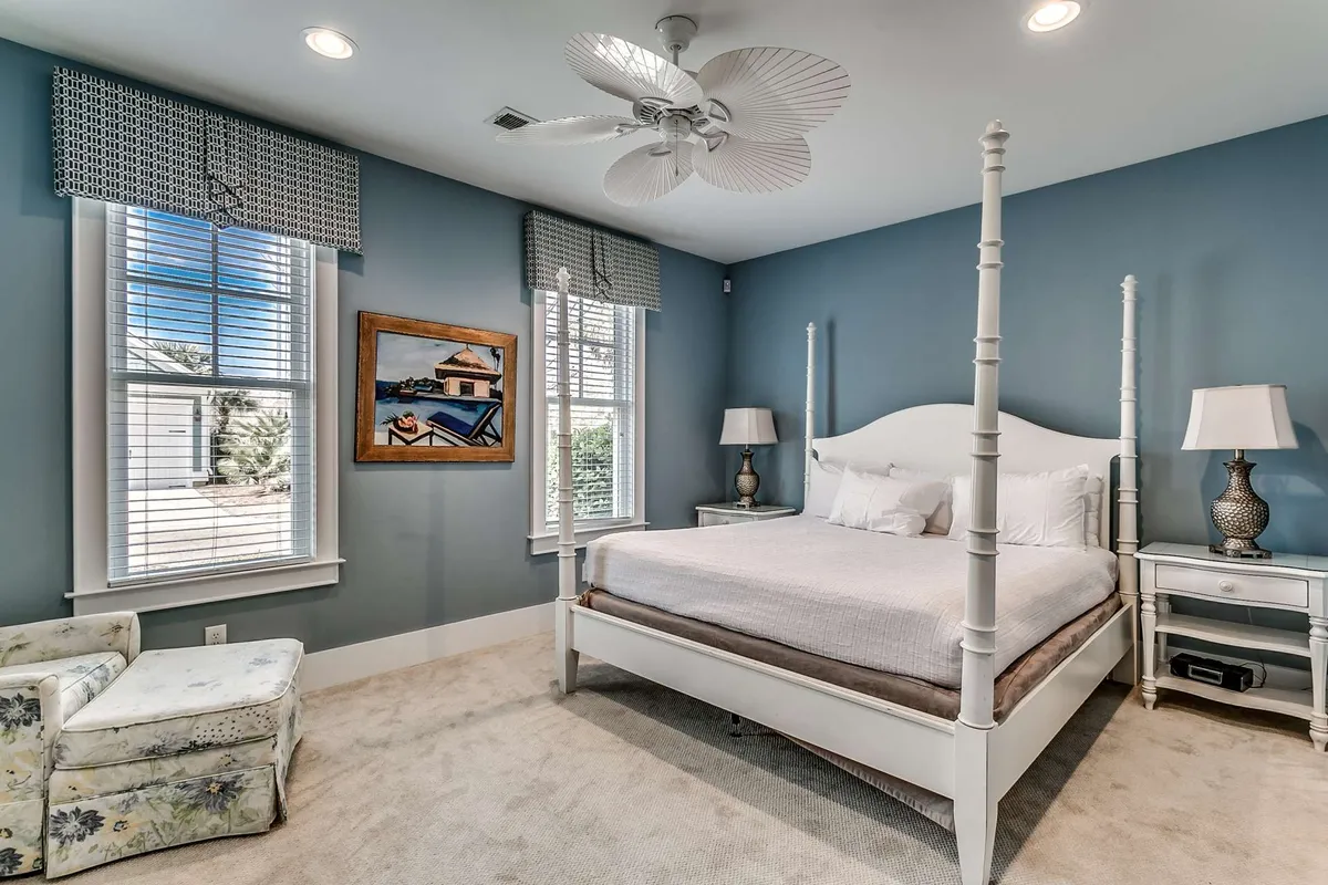 A serene bedroom scene featuring a four-poster bed dressed in soft linens, a framed painting on the wall, nightstand with a glowing lamp, and ample natural light streaming in through a window. The space feels tranquil and inviting, ideal for restful nights.
