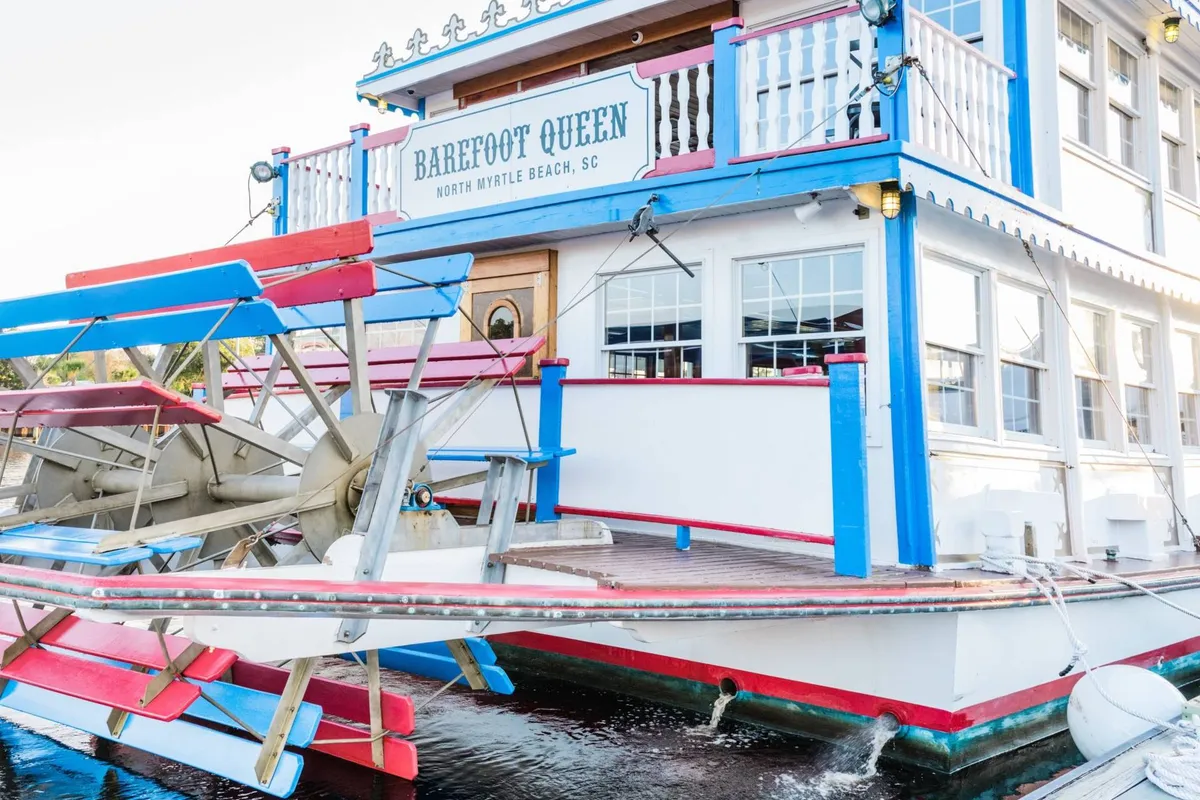 An exterior shot of a vibrant paddle wheel riverboat docked next to a colorful building facade, with clear signage indicating dining and entertainment options. The scene captures the festive mood with people visible on the deck, enjoying the view and atmosphere.