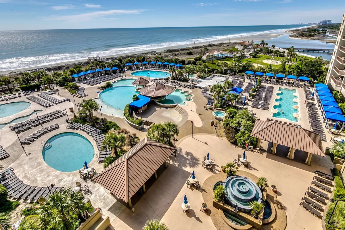 This image captures a vibrant resort pool area with multiple swimming pools, beachfront views, palm trees, lounge chairs, and inviting water features. The scene is lively, evoking leisure and vacation vibes, and illustrates guests enjoying the amenities under the sun.