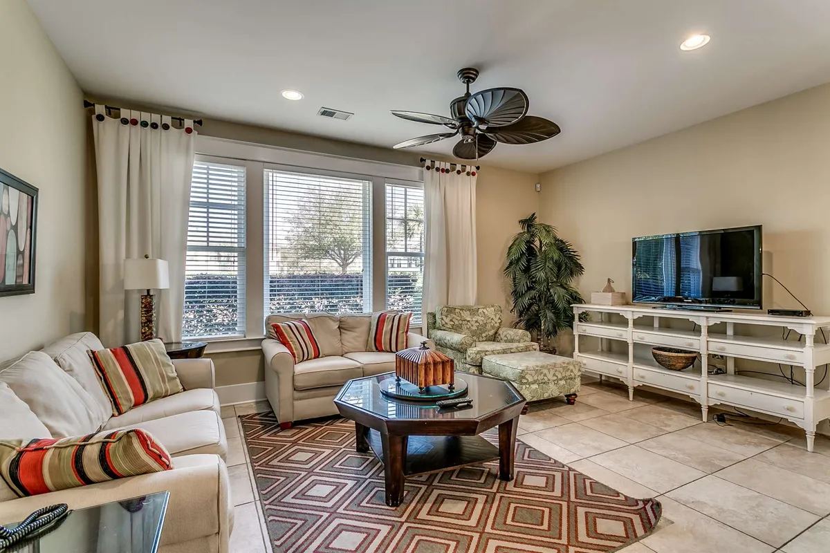 This image features a cozy living room with a sofa adorned with colorful cushions, a coffee table at the center, large windows that flood the room with natural light, a TV for entertainment, and indoor plants adding a homey touch. The scene conveys comfort and relaxation, perfect for gatherings or winding down after activities.