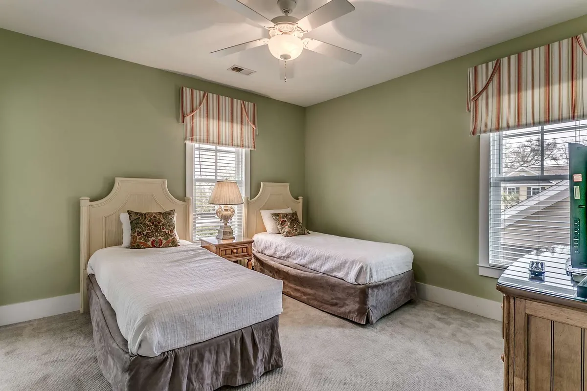 A twin bedroom with two neatly made beds, a decorative lamp on the nightstand, soft window treatments, and sunlight filtering in. The room feels quiet and inviting, suitable for children or additional guests seeking personal space.