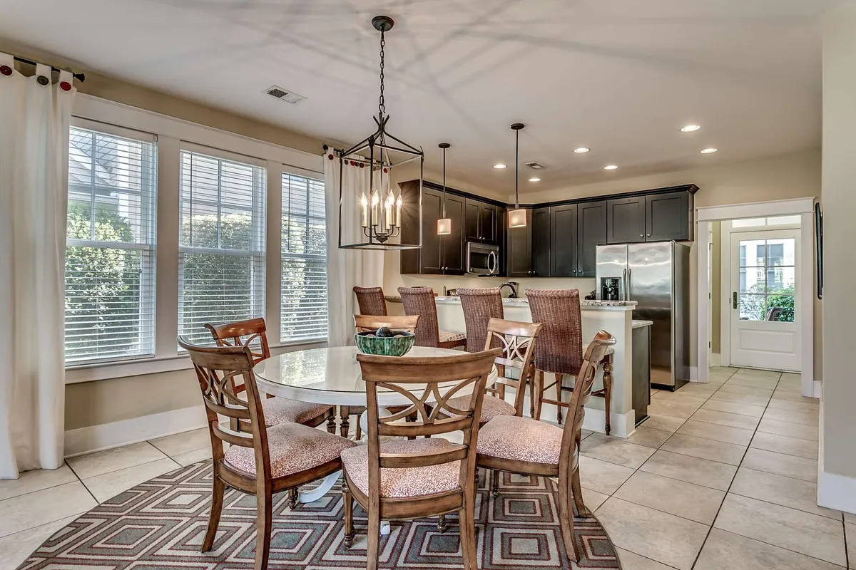 The image centers on a welcoming dining area, featuring a prominent dining table under a decorative chandelier, with a kitchen island visible in the background. The arrangement suggests a space ideal for family meals, group celebrations, and casual gatherings.