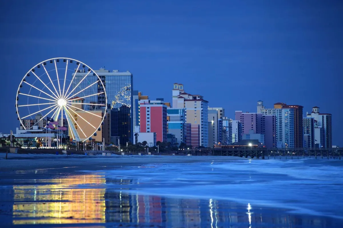 A lively exterior scene showing a Ferris wheel, colorful oceanfront buildings, and waves lapping the shore. The atmosphere is festive, with the lights and movement suggesting evening fun and exploration along the beach.