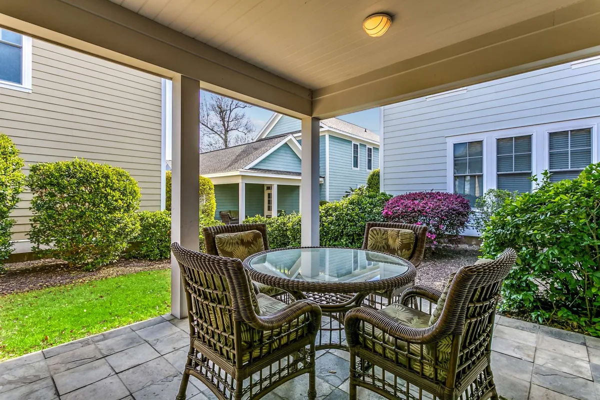 An exterior image of a covered patio furnished with a wicker dining set, surrounded by greenery. The tranquil outdoor atmosphere suggests the perfect setting for alfresco dining, morning coffee, or evening gatherings under the shelter of the patio.